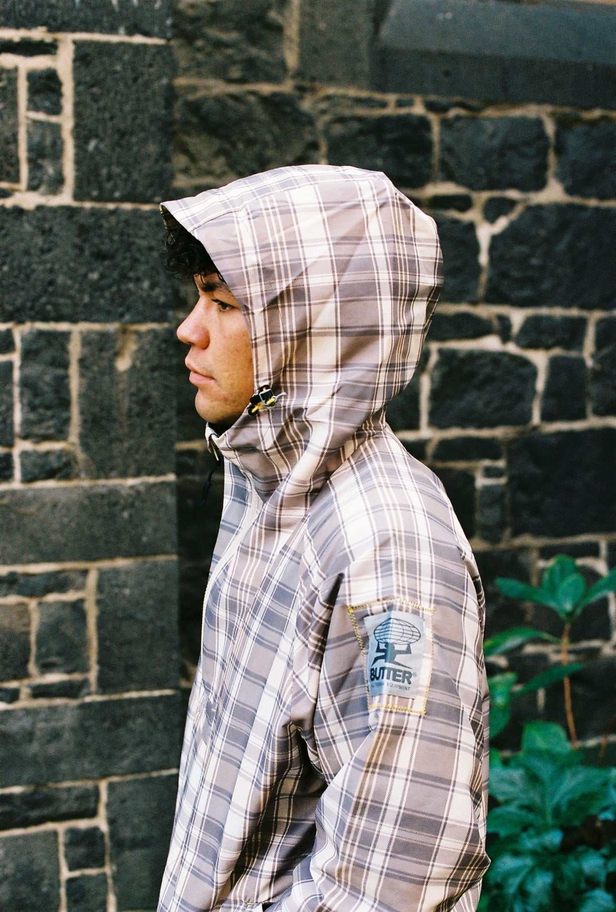 Person wearing a plaid jacket with a hood against a stone wall.