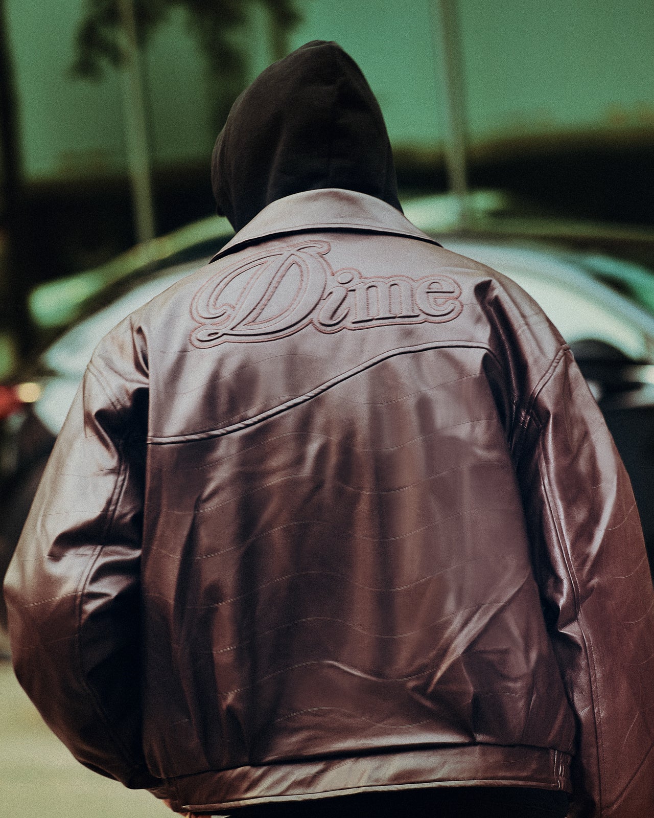 Person wearing a brown leather jacket with 'Dime' branding, standing in front of a blurred background.