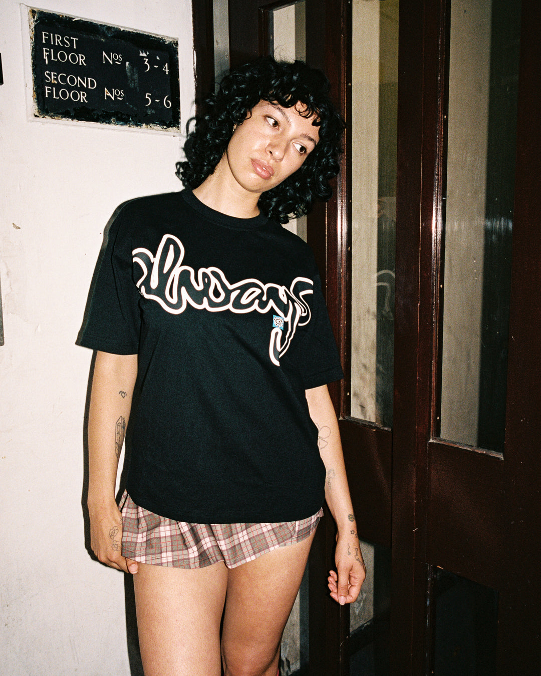 Person wearing a black t-shirt with white text and plaid shorts standing in front of a door.