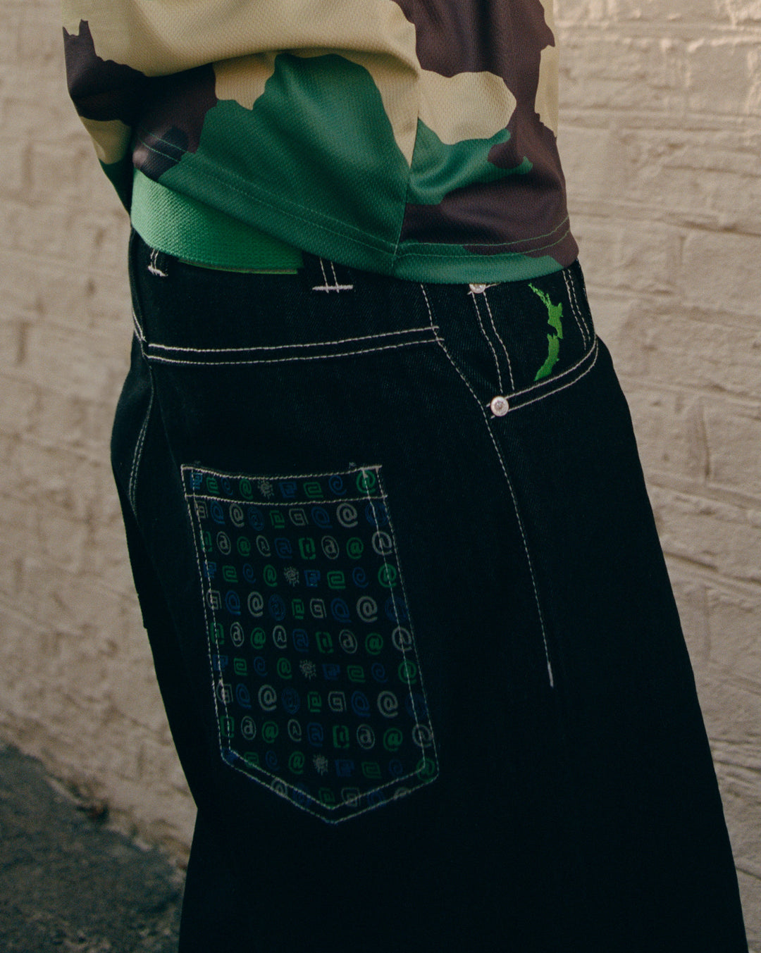 Close-up of black pants with a patterned pocket against a textured wall.