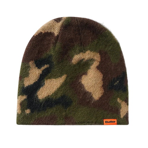 A knit beanie hat in a classic camouflage pattern of green, brown, black, and beige, featuring a small orange 'Butter' brand label on the lower right edge, set against a plain light gray background.