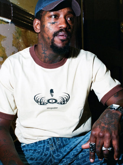 Man wearing a white t-shirt with a logo and blue cap, sitting indoors.