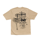 A beige Stussy short-sleeve t-shirt showing the back graphic print, which features a black illustration of a performer with a feathered hat, dice, and 'Los Angeles California USA' text.