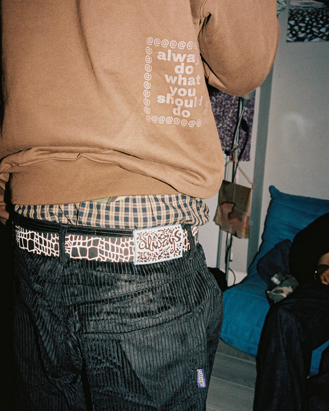 Person wearing a brown sweatshirt with text, plaid shirt, and patterned pants in an indoor setting.