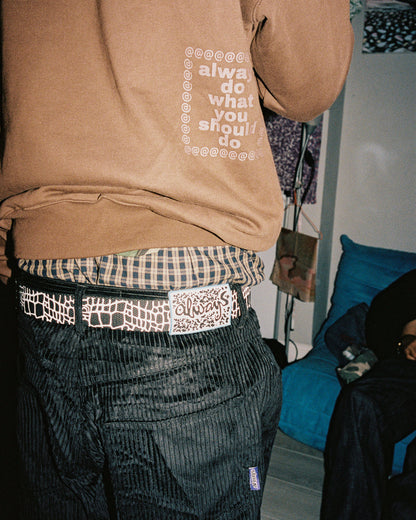 Person wearing a brown sweatshirt with text, plaid shirt, and patterned pants in an indoor setting.