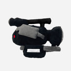 A plush toy shaped like a black video camera with gray details, a small red eye, and a white rectangular patch on the front, shown on a plain light gray background.