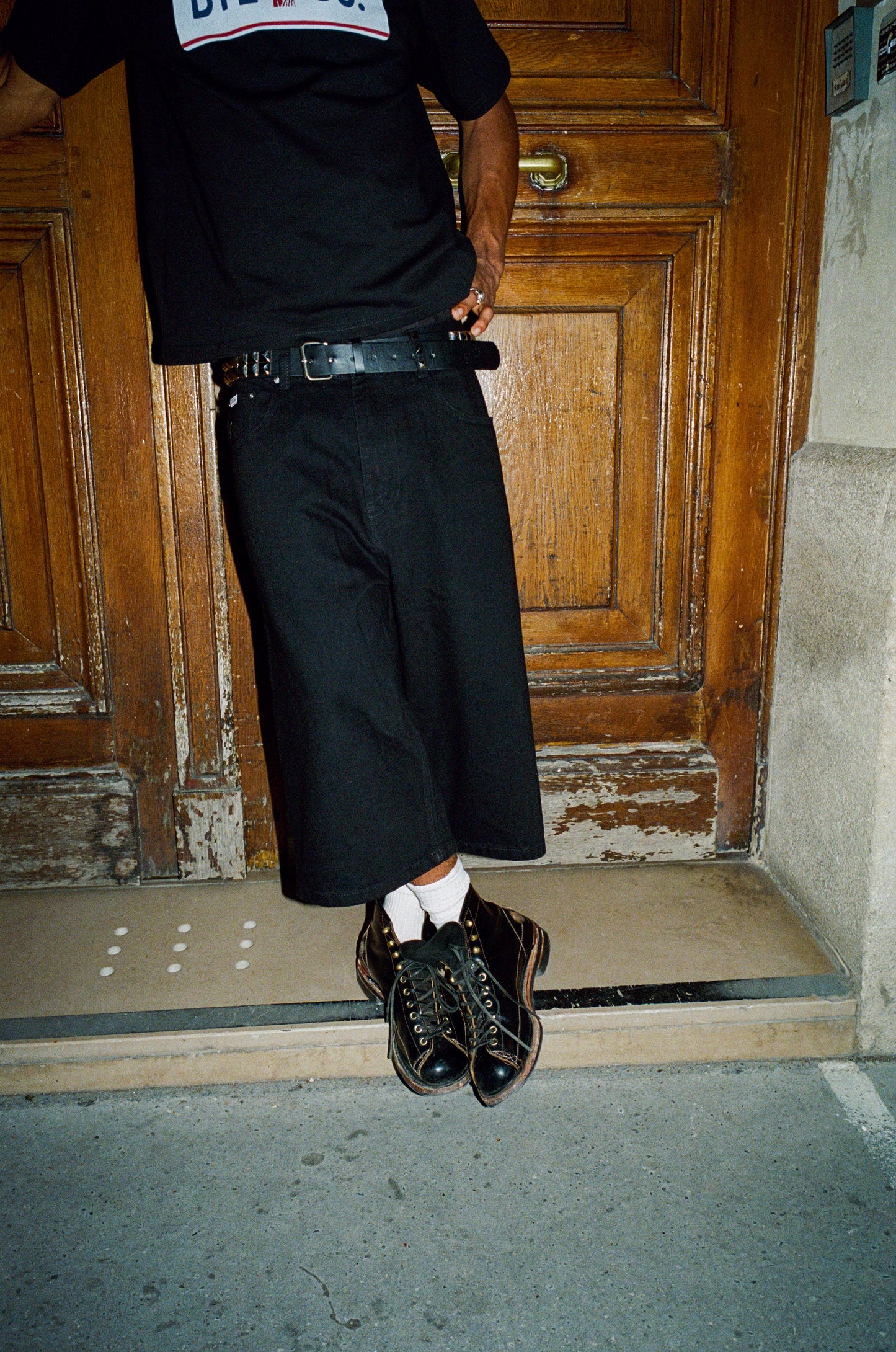 Person wearing a black outfit with a belt and shoes, standing in front of a wooden door.