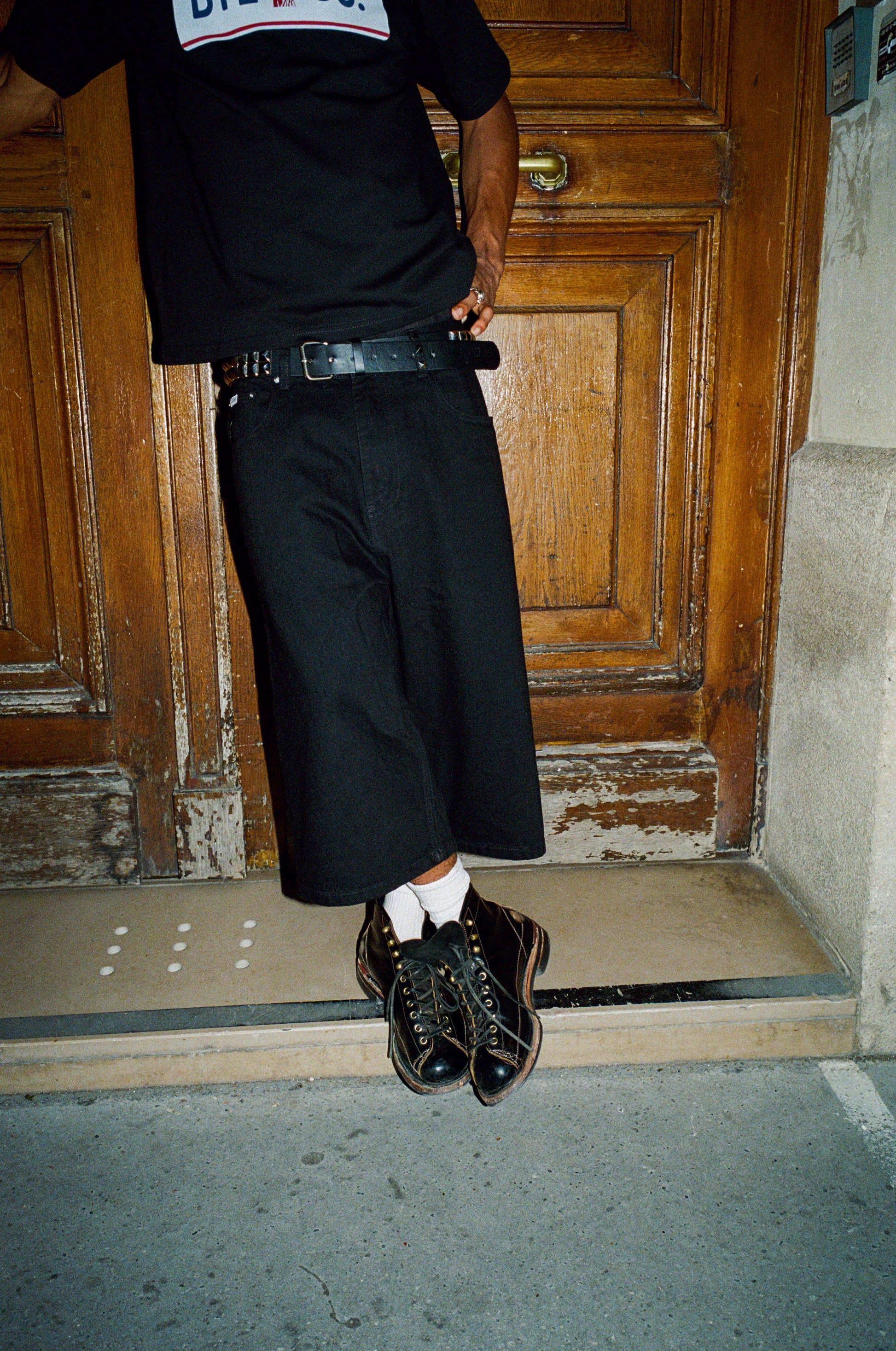 Person wearing a black outfit with a belt and shoes, standing in front of a wooden door.