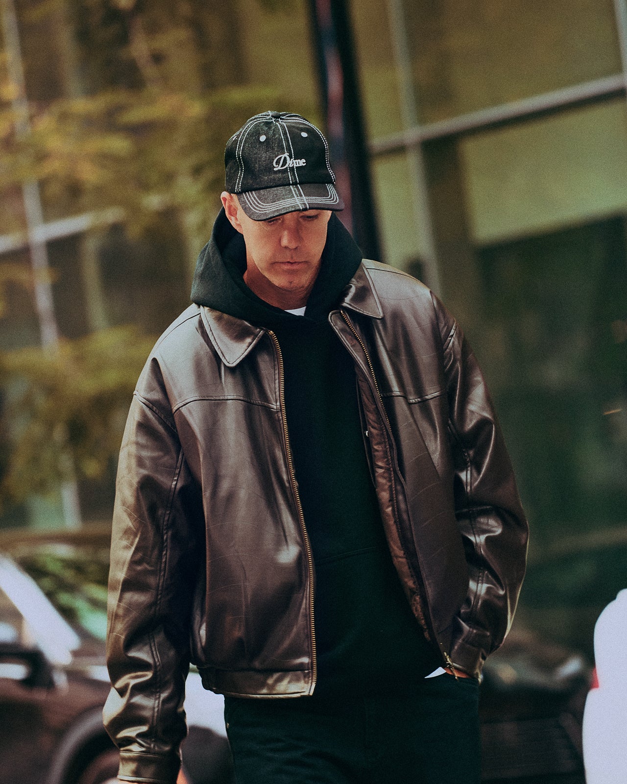 Man wearing a brown leather jacket and black cap walking outdoors.