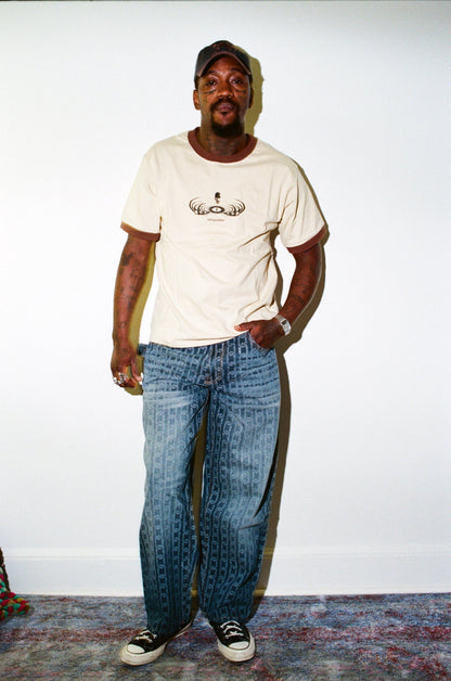 Man wearing a white t-shirt with a graphic design and blue jeans standing against a plain wall.